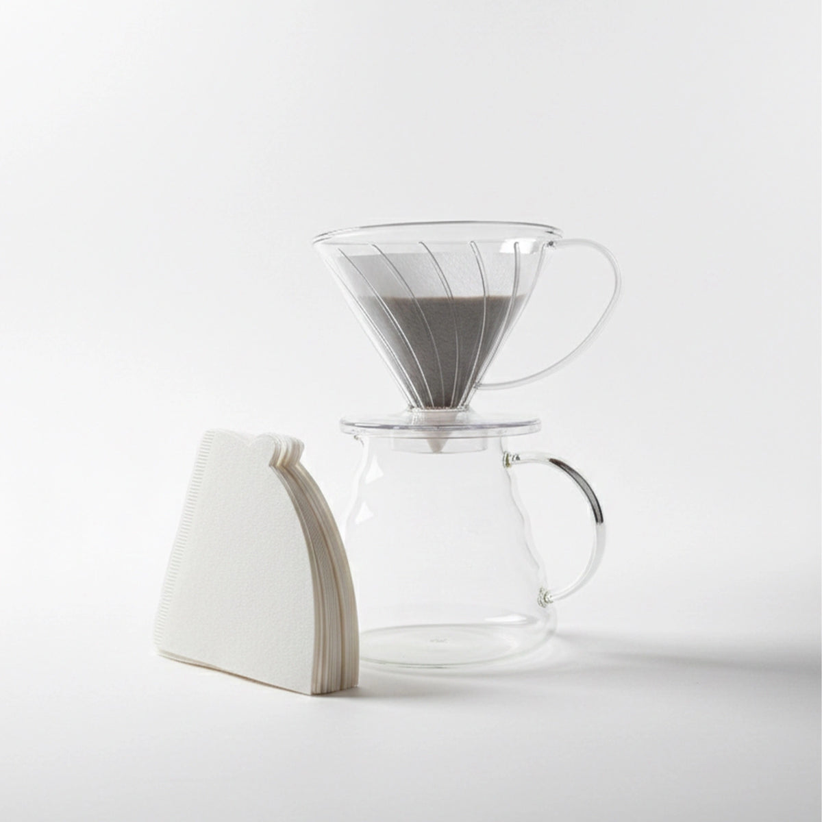 Kit de Preparo V60 02 montado: suporte transparente com filtro de papel sobre jarra de vidro, ao lado do maço de filtros